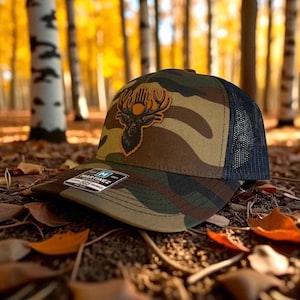 Op de afbeelding: Camouflage baseballpet met een hertenkoplogo. De pet heeft een bruin, groen en zwart camouflagepatroon met een donkerblauwe mesh-achterkant. Het hertenkoplogo is bruin en oranje. De pet ligt op een bed van bladeren.