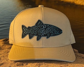 May include: A tan baseball cap with a curved peak, featuring a black and teal fish design. The fish is decorated with intricate patterns. The cap has a button on top. The design is reminiscent of tribal art.