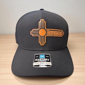 Può includere: Cappellino snapback nero con un design marrone e arancione con un simbolo a forma di croce e la parola "LUCERO". Il cappellino ha una visiera piatta e un'etichetta "Martinez Design Co.".