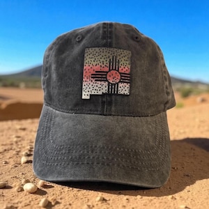 Può includere: Un berretto da baseball grigio scuro con una toppa colorata raffigurante il contorno dello stato del New Mexico e un simbolo Zia. La toppa include un design arcobaleno e un motivo a pois. Il berretto è appoggiato su una superficie sabbiosa.