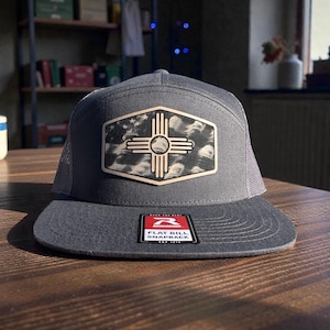 Op de afbeelding: Een grijze flat bill snapback pet met een mesh achterkant. De voorkant heeft een patch met een zuidwestelijk ontwerp in zwart, wit en bruin. De pet heeft een platte klep en een label met "FLAT BILL SNAPBACK".