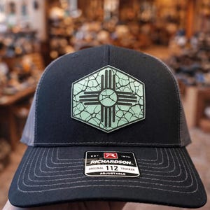 May include: Black trucker hat with a gray mesh back and a patch featuring the New Mexico Zia symbol in light green. The hat has a curved brim and an adjustable strap. The Richardson brand label is visible.