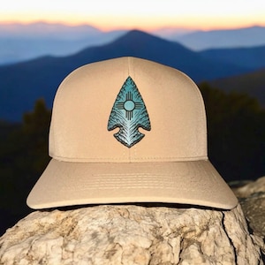 Puede incluir: Gorra de béisbol color canela con un diseño de punta de flecha turquesa y un símbolo de sol. La gorra está colocada contra un telón de fondo de montañas y una puesta de sol, mostrando un tema del suroeste.