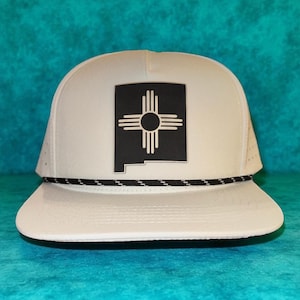 Op de afbeelding: Witte truckerpet met een zwart-wit New Mexico Zia-symbool. De pet heeft een gebogen klep, een zwart-wit touwdetail en een mesh achterkant. Een klassiek accessoire.