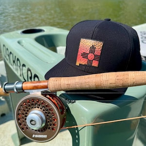 Può includere: Un cappellino da baseball nero con una toppa con la bandiera dello stato del New Mexico, una canna da pesca e un mulinello. La canna da pesca ha un manico in sughero e si trova su una superficie verde. Il mulinello è color bronzo.