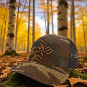 Op de afbeelding: Camouflage baseballpet met een bruin leren embleem met een gestileerd Zia-symbool en een silhouet van een hert. De pet heeft een mesh rug en een gebogen klep. De achtergrond toont een bos met gele bladeren.