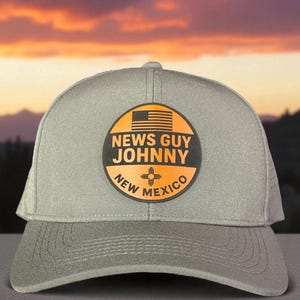 May include: Gray trucker hat with a circular patch featuring the text "NEWS GUY JOHNNY" and "NEW MEXICO". The patch includes an American flag and a New Mexico Zia symbol. The hat has a curved brim and mesh back.