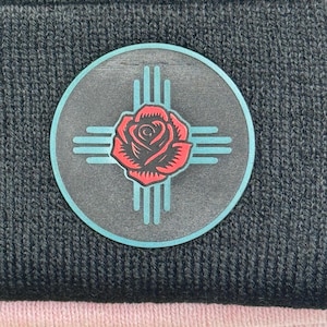 Op de afbeelding: Een donkergrijze beanie met een cirkelvormig embleem. Het embleem heeft een rode roos, turquoise lijnen en een turquoise rand. De beanie is gemaakt van gebreid materiaal.