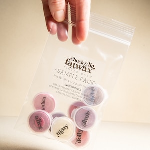 May include: A clear plastic sample pack of Cheek & Lip Fatwax tinted balm. The pack contains several small, round containers of balm in various shades, each labeled with a name. The text "SAMPLE PACK" is visible on the packaging. The net weight is .07 oz / 2 g per clamshell.