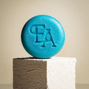 May include: A teal blue round soap bar with the letters "EA" embossed on the top. The soap bar is sitting on a white and brown textured surface.