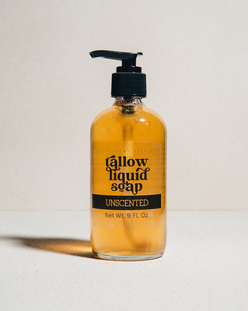 May include: A clear amber glass bottle with a black pump dispenser filled with a yellow liquid. The bottle has a black label with the text "tallow liquid soap" and "UNSCENTED" in a retro font. The label also states "Net Wt. 9 Fl. Oz."
