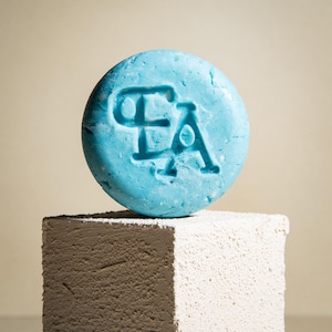 May include: A light blue solid shampoo bar with the letters "EA" embossed on the top. The bar is sitting on a white and gray textured surface.