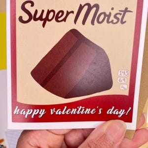 May include: A Valentine's Day card with a red background and the text "Super Moist" in a vintage style font. A chocolate bar is pictured in the center of the card. The text "happy valentine's day!" is printed in white at the bottom of the card.