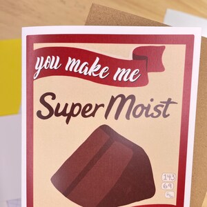 May include: A red and white card with the text "you make me Super Moist" and a brown chocolate bar illustration. The card has a red ribbon with the text "Super Moist" printed on it.