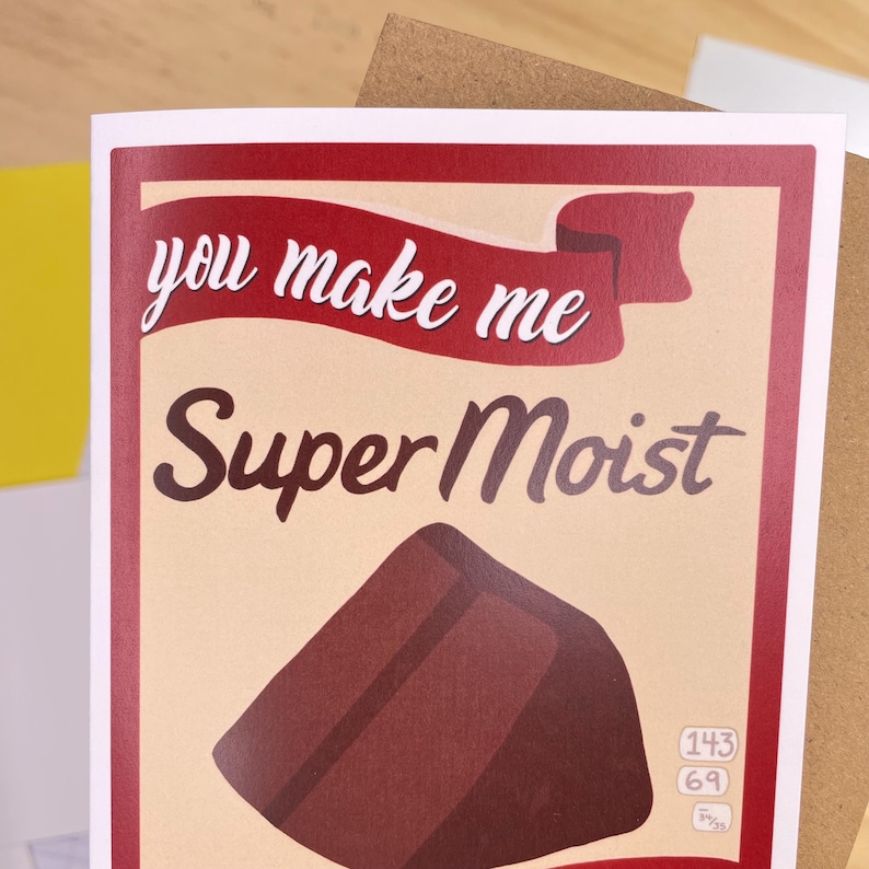 May include: A red and white card with the text "you make me Super Moist" and a brown chocolate bar illustration. The card has a red ribbon with the text "Super Moist" printed on it.