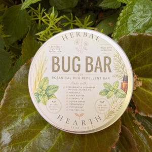 May include: A round metal tin of Herbal Bug Bar botanical bug repellent, weighing 2.5 oz or 70 g. The label features illustrations of herbs and lists ingredients including peppermint, spearmint, jojoba oil, beeswax, shea butter, and citronella.