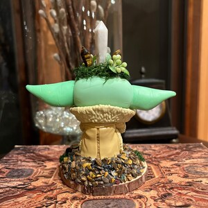 Puede incluir: Figura verde claro de un personaje con orejas grandes, que lleva un abrigo color canela con cuello de piel. La figura tiene una cabeza verde con un peque&ntilde;o cristal y plantas artificiales encima. La base est&aacute; decorada con piedras peque&ntilde;as.