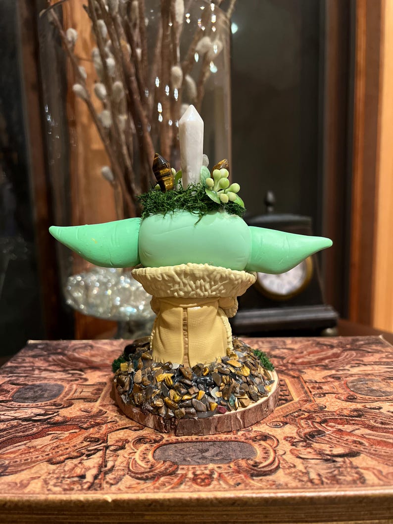 Puede incluir: Figura verde claro de un personaje con orejas grandes, que lleva un abrigo color canela con cuello de piel. La figura tiene una cabeza verde con un peque&ntilde;o cristal y plantas artificiales encima. La base est&aacute; decorada con piedras peque&ntilde;as.