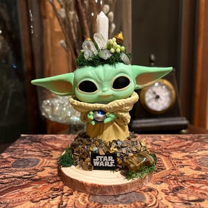 Puede incluir: Una figura verde de un personaje de Star Wars, con grandes orejas y ojos negros. La figura est&aacute; adornada con cristales y musgo, y se encuentra sobre una base con el logotipo de Star Wars.