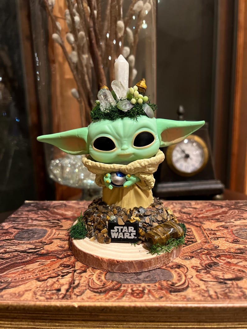 Puede incluir: Una figura verde de un personaje de Star Wars, con grandes orejas y ojos negros. La figura est&aacute; adornada con cristales y musgo, y se encuentra sobre una base con el logotipo de Star Wars.