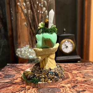 Puede incluir: Una escultura decorativa con una forma redondeada verde claro, coronada por un cristal y peque&ntilde;os elementos decorativos. La escultura descansa sobre una base adornada con peque&ntilde;as piedras y vegetaci&oacute;n. La base est&aacute; sobre un libro con un dise&ntilde;o ornamentado.