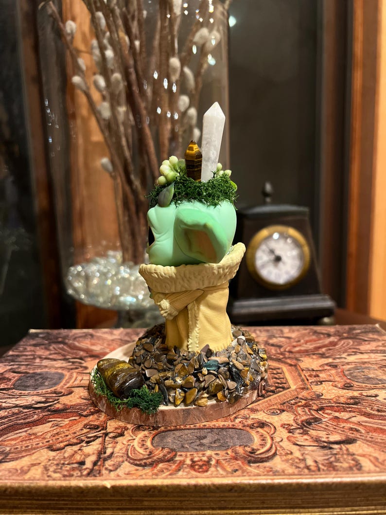 Puede incluir: Una escultura decorativa con una forma redondeada verde claro, coronada por un cristal y peque&ntilde;os elementos decorativos. La escultura descansa sobre una base adornada con peque&ntilde;as piedras y vegetaci&oacute;n. La base est&aacute; sobre un libro con un dise&ntilde;o ornamentado.