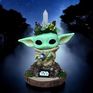 Puede incluir: Una figura verde de un personaje con orejas grandes, sosteniendo una esfera plateada. La figura est&aacute; adornada con cristales y vegetaci&oacute;n, y se encuentra sobre una base de madera con el logo "STAR WARS". El fondo presenta un cielo estrellado.
