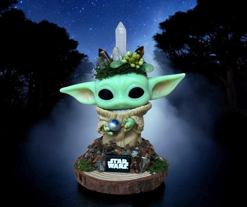 Puede incluir: Una figura verde de un personaje con orejas grandes, sosteniendo una esfera plateada. La figura est&aacute; adornada con cristales y vegetaci&oacute;n, y se encuentra sobre una base de madera con el logo "STAR WARS". El fondo presenta un cielo estrellado.