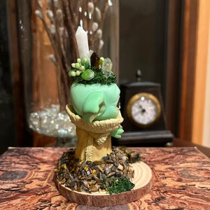 Puede incluir: Una figura verde con una cabeza verde claro, adornada con cristales y una vela blanca. La figura est&aacute; sobre una base de madera cubierta de peque&ntilde;as piedras y se asienta sobre un libro decorativo. Un jarr&oacute;n de cristal con flores blancas est&aacute; en el fondo.