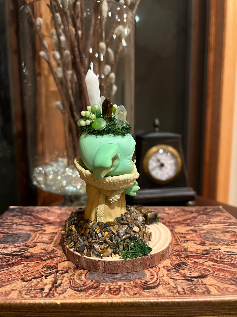 Puede incluir: Una figura verde con una cabeza verde claro, adornada con cristales y una vela blanca. La figura est&aacute; sobre una base de madera cubierta de peque&ntilde;as piedras y se asienta sobre un libro decorativo. Un jarr&oacute;n de cristal con flores blancas est&aacute; en el fondo.