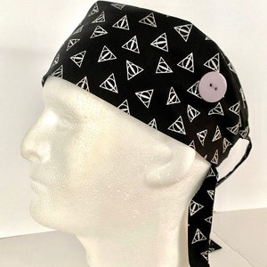 May include: Black surgical scrub hat with a white repeating pattern of the Deathly Hallows symbol. The hat has a button on the side for attaching a face mask.