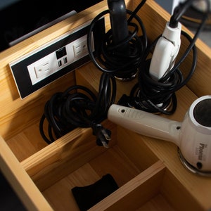 May include: A wooden drawer with compartments for storing cords and electrical devices. The drawer contains a hair dryer, a flat iron, and a power strip with multiple outlets.