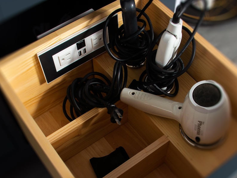 May include: A wooden drawer with compartments for storing cords and electrical devices. The drawer contains a hair dryer, a flat iron, and a power strip with multiple outlets.