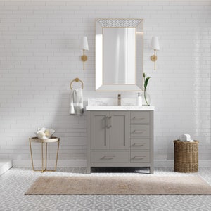 May include: A grey bathroom vanity with two doors and three drawers. The vanity has a white countertop and a gold tap. The vanity is in front of a white tiled wall with a gold mirror above it.