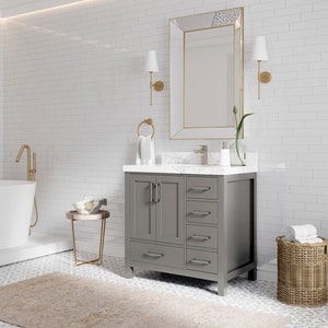 May include: A grey bathroom vanity with a white countertop and a white porcelain sink. The vanity has four drawers and two doors. The vanity is in front of a white subway tile wall. There is a gold-framed mirror above the vanity and a gold faucet.