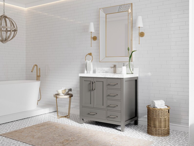 May include: A grey bathroom vanity with a white countertop and a white porcelain sink. The vanity has four drawers and two doors. The vanity is in front of a white subway tile wall. There is a gold-framed mirror above the vanity and a gold faucet.