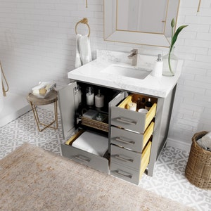 May include: A grey bathroom vanity with a white countertop and a white porcelain sink. The vanity has four drawers and a cabinet door. The drawers are open, revealing wooden drawer boxes. The vanity is in a bathroom with white subway tile walls and a patterned floor.