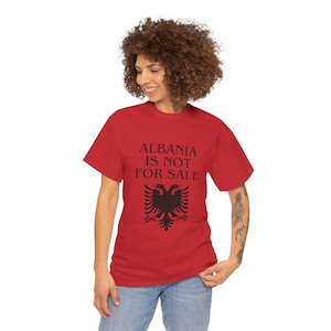 May include: A red short-sleeved t-shirt featuring the black text ALBANIA IS NOT FOR SALE printed on the chest above a black graphic of a double-headed eagle, which is the national emblem of Albania, centered on the front of the garment.