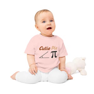 Peut inclure: Un t-shirt pour bébé à manches courtes rose clair avec le texte marron Cutie Pie au-dessus d'un symbole d'angle mathématique noir et du symbole pi. Le t-shirt a une encolure ronde et est porté avec un legging blanc uni, formant une tenue simple.