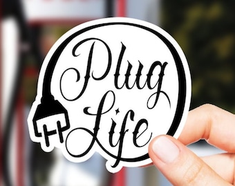 May include: A black and white sticker with the text "Plug Life" in a circle with a stylized plug icon. The sticker is being held in front of a Tesla charging station.