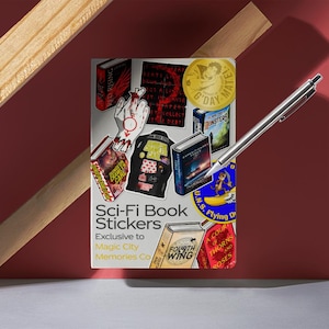 May include: A collection of Sci-Fi Book Stickers featuring various book covers and graphics. The stickers are exclusive to Magic City Memories Co. A silver pen rests on the stickers, with a red background and wooden accents.