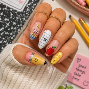 May include: A set of almond-shaped press-on nails featuring school-themed designs. The nail art includes a yellow pencil tip, a stack of books with a smiley face, white lined paper with a heart, a red apple, and a yellow floral pattern on a nude base.