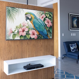 May include: A large, framed artwork of a blue, yellow, and green macaw parrot perched among pink hibiscus flowers. The artwork is mounted on a wooden wall, above a white shelf with a black media device.