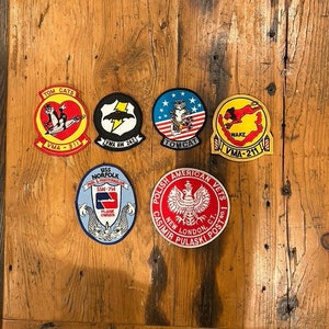 Puede incluir: Una colección de parches bordados coloridos con aviones militares y nombres de unidades, incluyendo "Tom Cats", "VMA-311", "VMA AW 242", "TOMCAT", "WAKE", "VMA-211", "USS NORFOLK SSN-714", "PLANK OWNER", y "POLISH AMERICAN VETS CASIMIR PULASKI POST NO.1 NEW LONDON, CT."