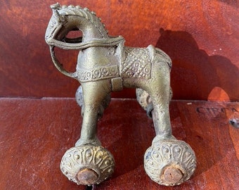 May include: A gold-colored metal toy horse with wheels. The horse has intricate details and a saddle. The wheels are round and have a decorative design.
