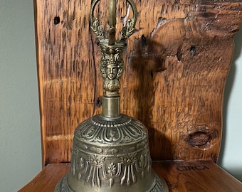 May include: A brass bell with intricate carvings and a decorative handle. The bell is sitting on a wooden surface.