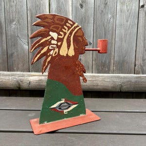 May include: A metal sculpture of a Native American man wearing a headdress and smoking a pipe. The sculpture is painted in shades of brown, green, and white. The base of the sculpture is a flat, rectangular piece of metal.