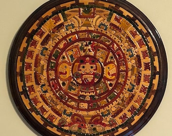 May include: A round, wooden wall hanging with a colorful depiction of the Aztec calendar, also known as the Sun Stone. The calendar features a central face surrounded by concentric circles of glyphs and symbols.