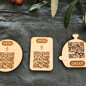 May include: Three wooden coasters with QR codes and the words "MENU" and "ORDER" printed on them. The coasters are on a gray fabric surface.