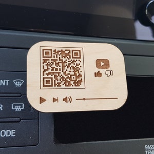 May include: A wooden car air freshener with a QR code, YouTube play button, thumbs up and down icons, and play, rewind, and volume control icons. The air freshener is attached to a car's dashboard.